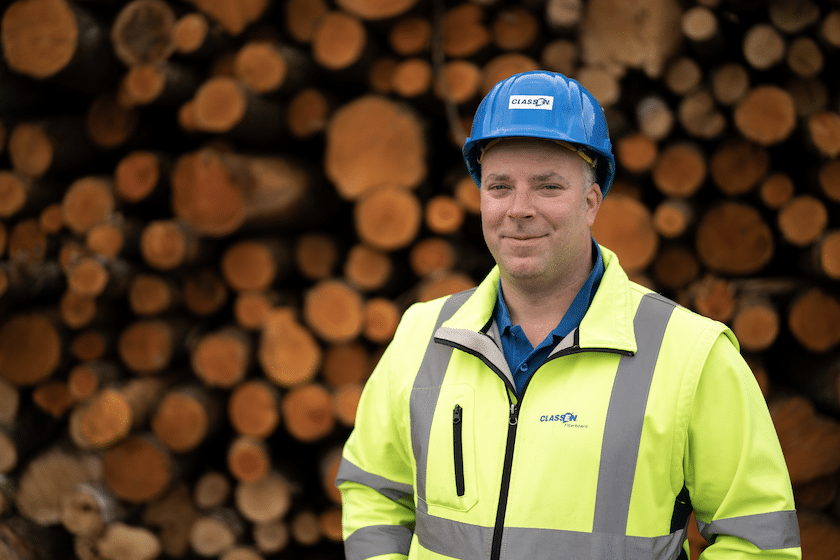 Ein CLASSEN Mitarbeiter mit Schutzhelm und Warnweste steht vor einem großen Stapel frisch geschnittener Holzstämme. Das Bild symbolisiert die nachhaltige Holzverarbeitung und die Rohstoffbasis für innovative Bodenbeläge von CLASSEN