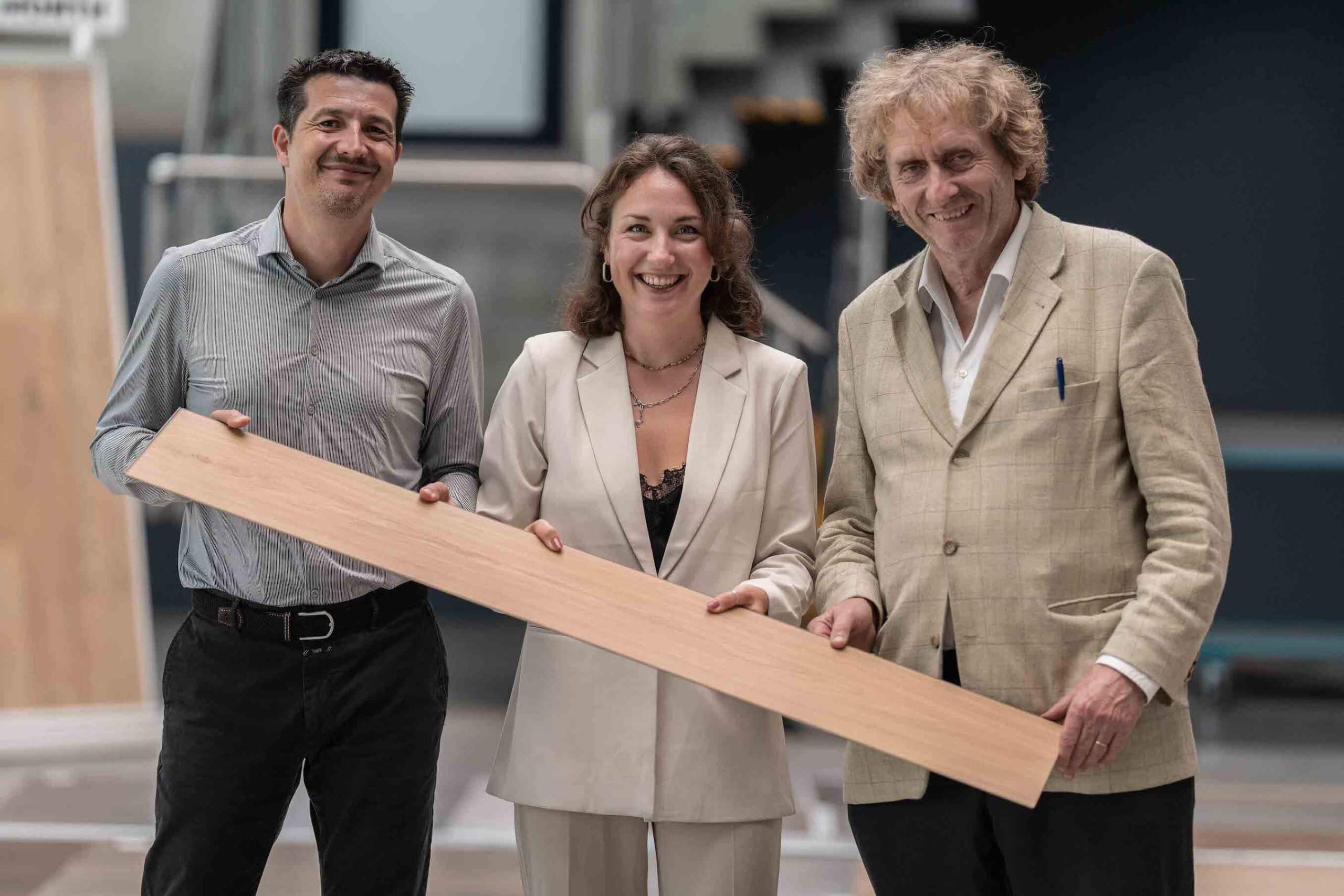 Drei Personen halten gemeinsam ein langes Bodenpaneel in den Händen und lächeln in die Kamera – Symbol für Teamarbeit, Innovation und die nachhaltige Entwicklung von Bodenbelägen bei CLASSEN.