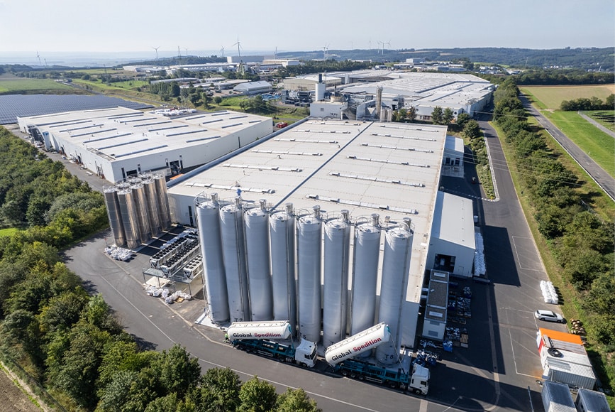 Luftaufnahme des CLASSEN Werks mit großen Industriesilos, Produktionshallen und angrenzender Infrastruktur, umgeben von grüner Landschaft und Windrädern – Symbol für moderne, nachhaltige und ressourcenschonende Bodenbelagsproduktion in Deutschland