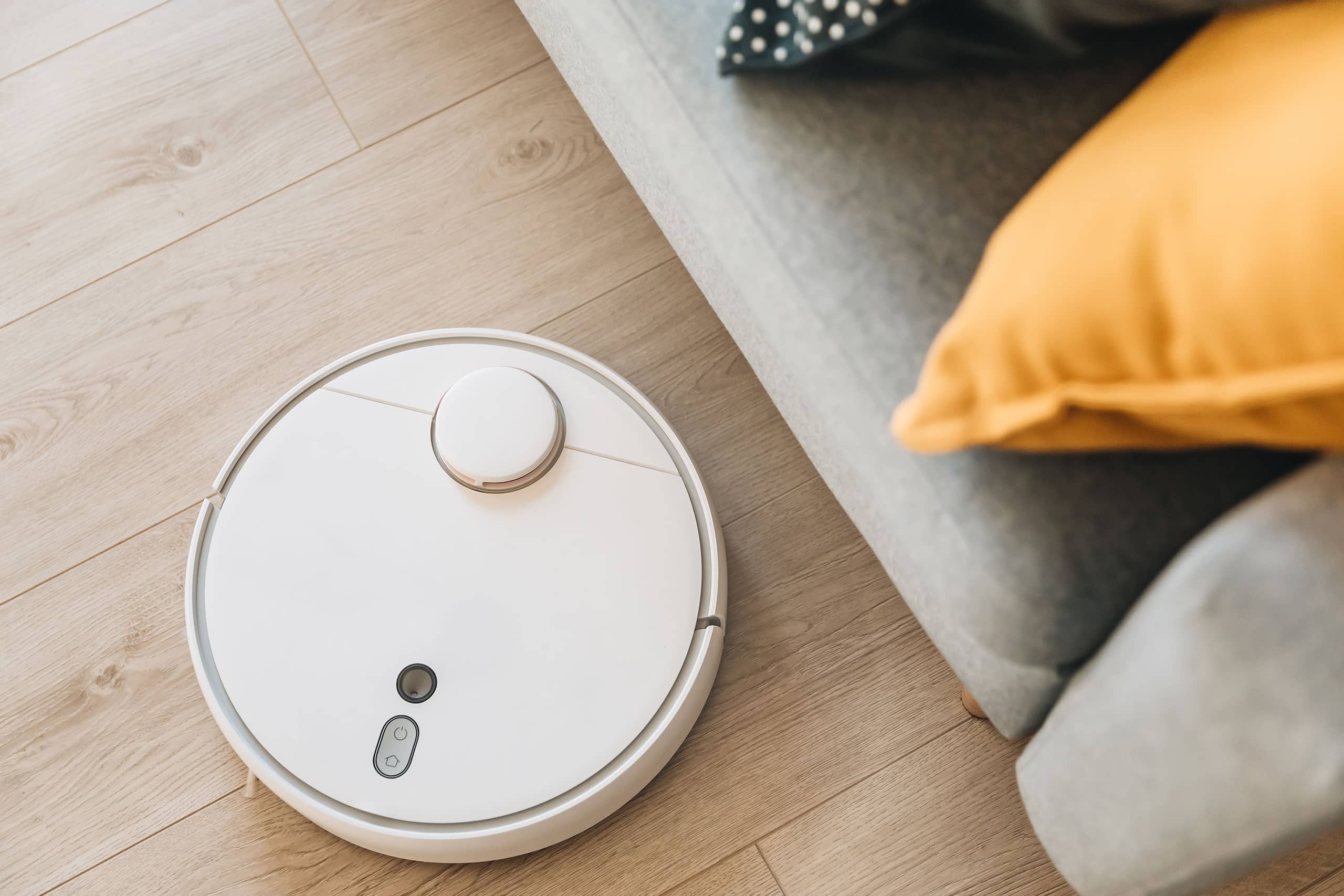 Weißer Saugroboter fährt über hellen Holzoptik-Designboden neben grauem Sofa mit orangenem Kissen.
